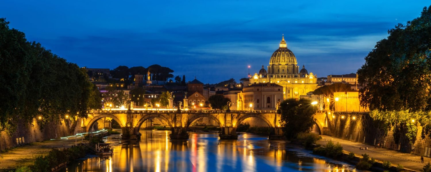 View of the Tiber River in the center of Rome at night, Italy. Embankment street, multiple buildings and Saint Peter Basilica in the distance